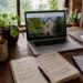 Laptop mit geöffneter Webseite, Notizbuch und Handy auf einem Holztisch am Fenster.
